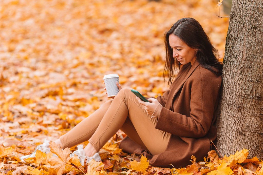 Fall concept - beautiful woman drinking coffee in autumn park under fall foliage