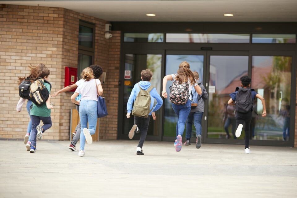 Group Of High School Students Running Into School Building At Beginning Of Class