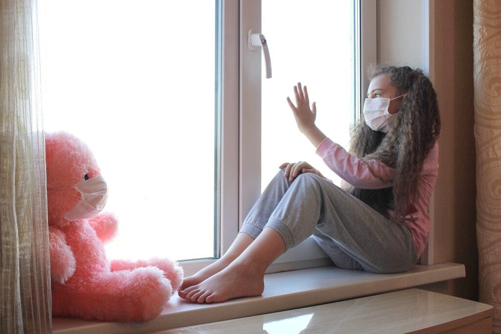 A girl in a medical mask sits on the windowsill and touches the window with her hand
