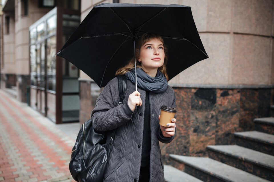 Young cute lady standing on street with black umbrella and coffee in hands