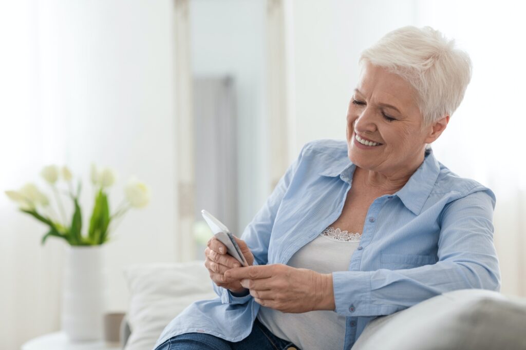Happy elderly lady using mobile phone while relaxing on couch at home