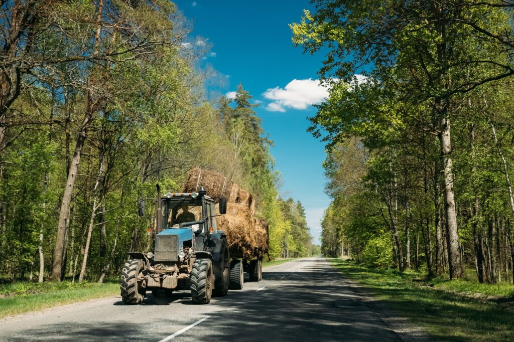 Tractor Is Carrying Hay On Cart. Tractor On Country Road Through
