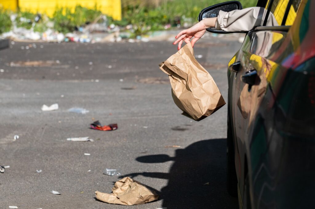 Woman holding trash paper bag in hand, out of window driving in car.