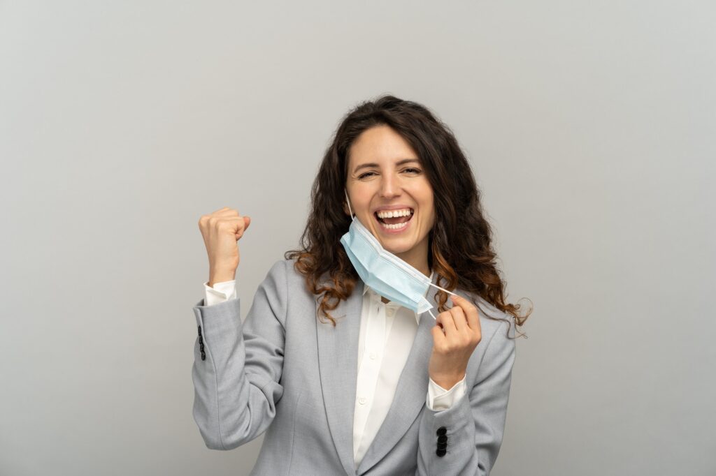 Studio portrait of happy business woman taking off mask from face, raising fist on grey background.
