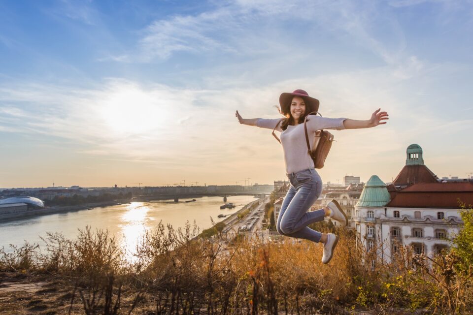 Young student woman jumping opposite famous facade and entrance to Hotel Gellert on banks of Danube