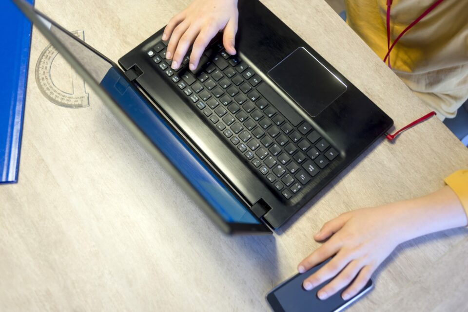Hands of a young boy typing on a laptop keyboard over a rustic wooden table.